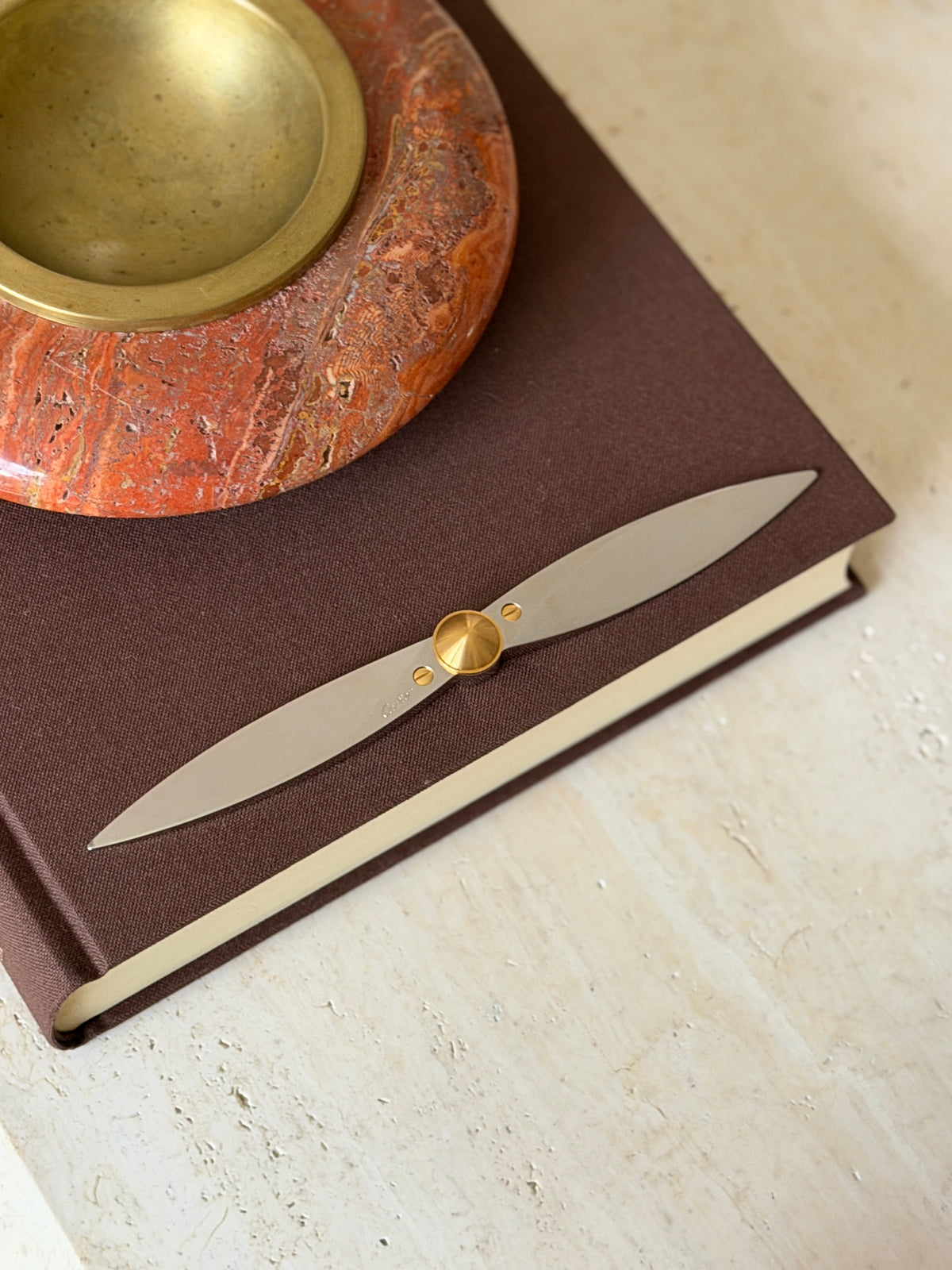Cartier propeller letter opener laid across a brown clothbound book beside a red stone ashtray with a brass well, steel blades meeting at a gold cone hub