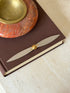 Cartier propeller letter opener laid across a brown clothbound book beside a red stone ashtray with a brass well, steel blades meeting at a gold cone hub