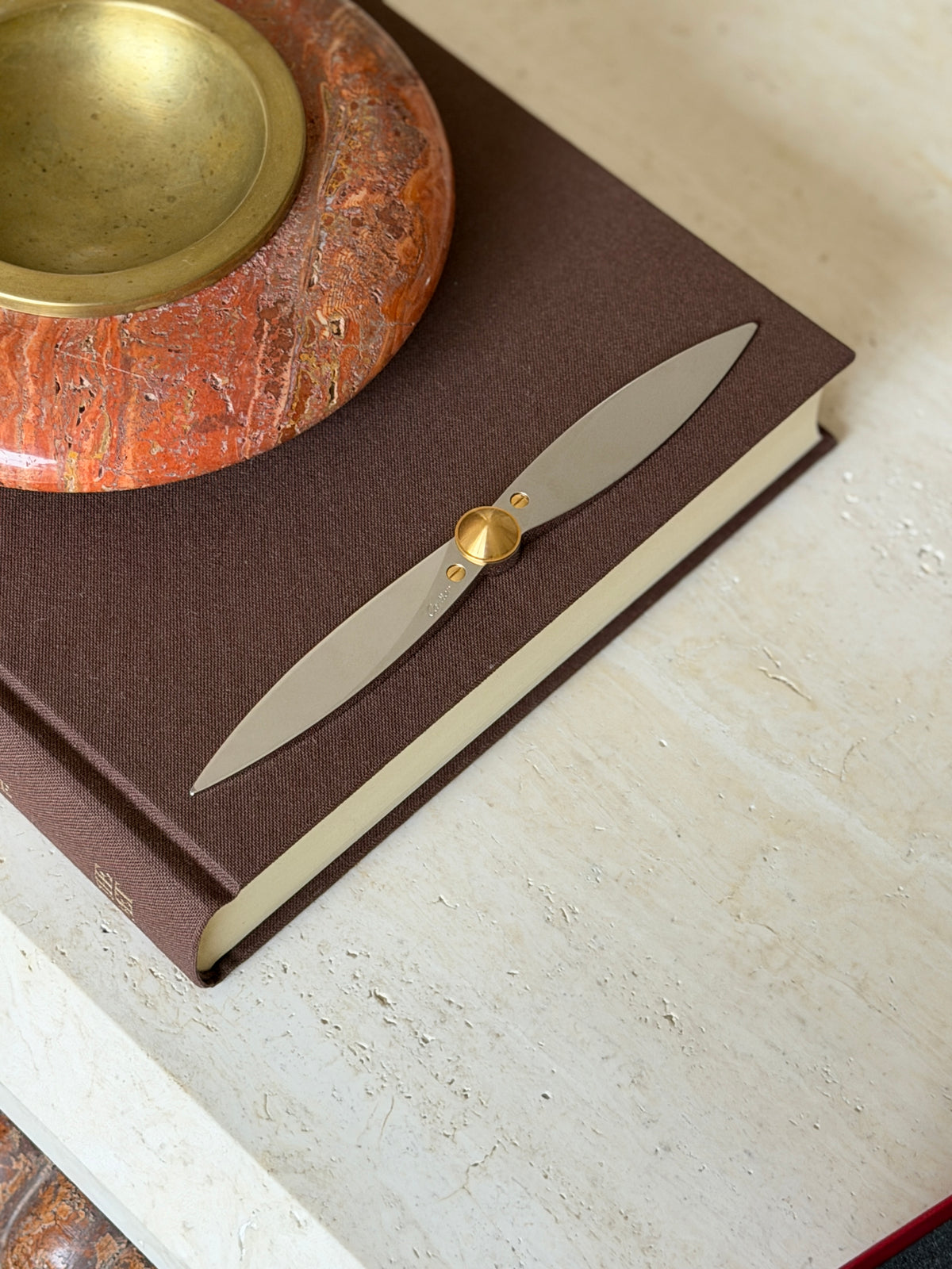 Cartier letter opener shaped like an aircraft propeller, styled on a brown book near a red stone ashtray with the edge of the red box in view