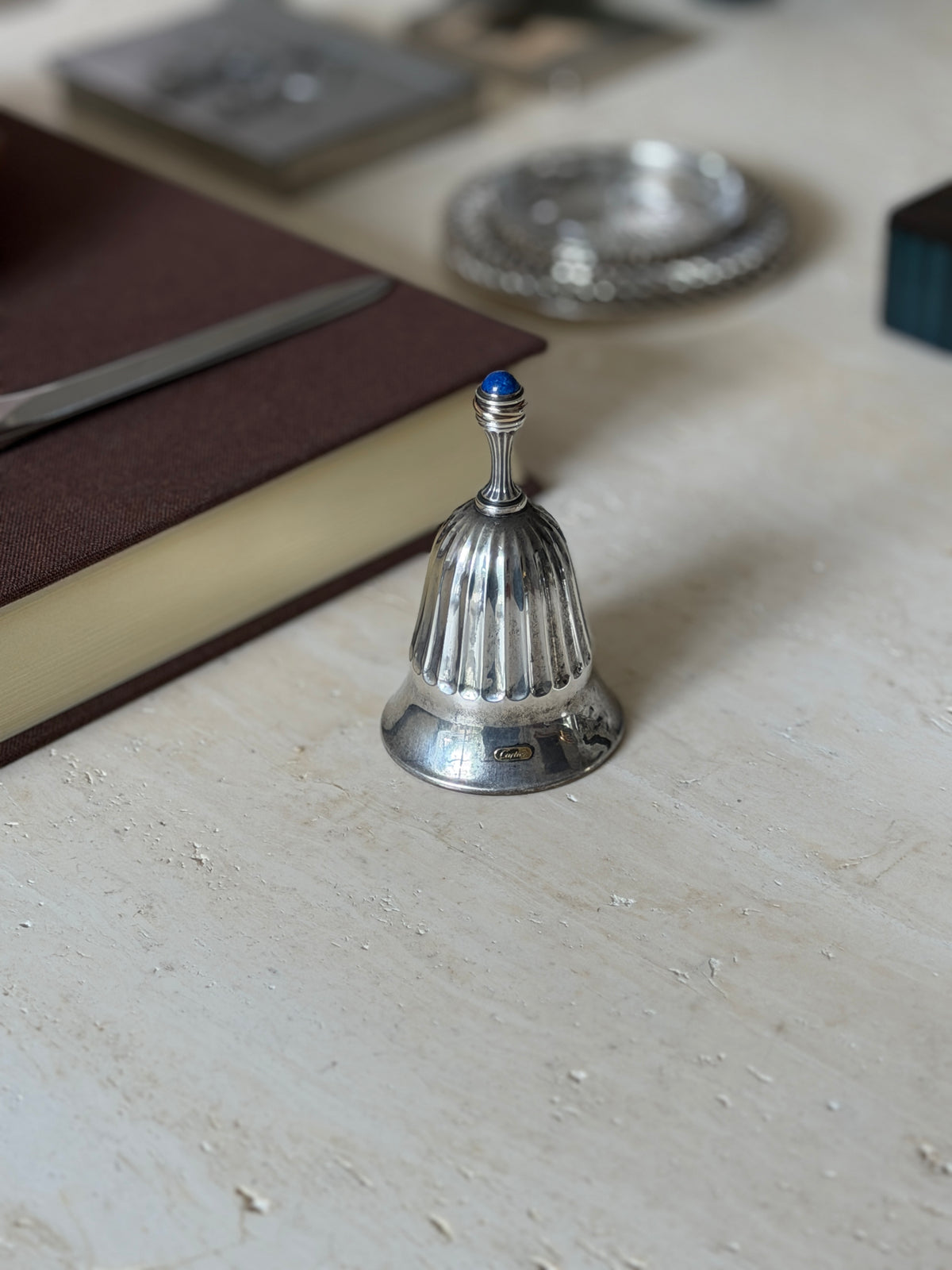 The bell viewed with a letter opener resting on the book edge and a woven silver coaster blurred behind