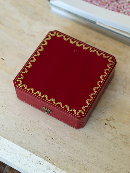 Cartier red leather presentation box closed, with gold tooled scrollwork border and brass clasp, resting on a stone surface