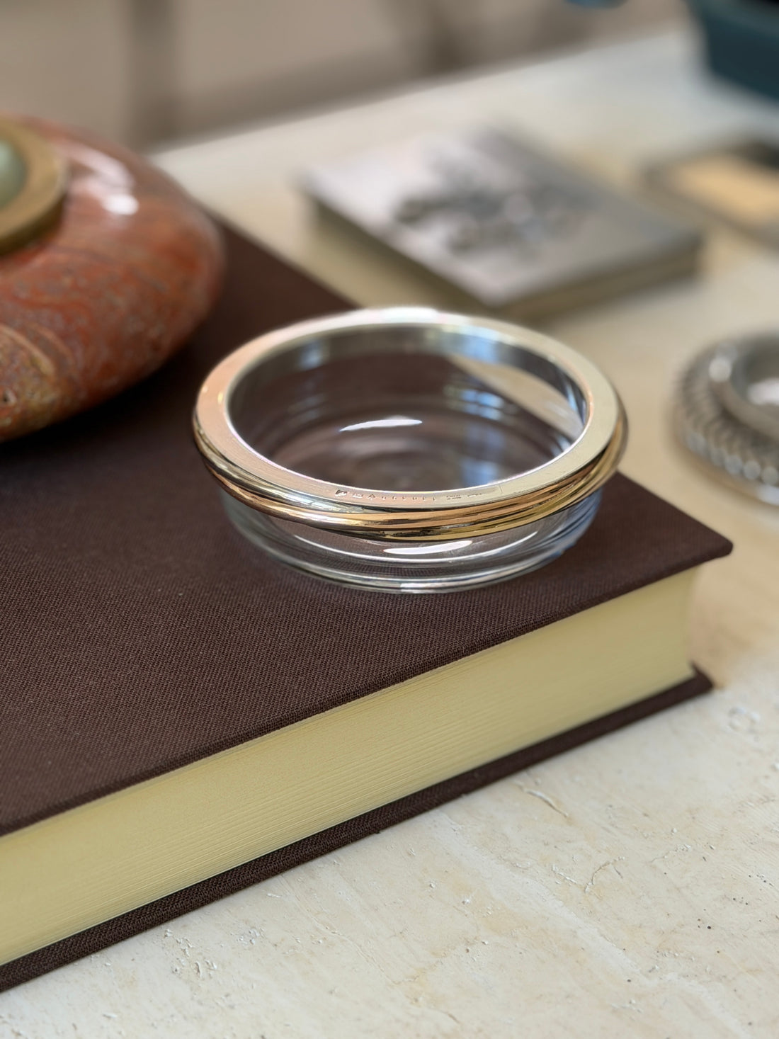 Cartier Trinity crystal dish displayed on a book alongside other tabletop objects, including a stone catchall and a silver rimmed dish