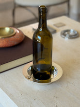 Cartier bottle coaster holding a green wine bottle on a travertine table beside a brown book and red stone ashtray