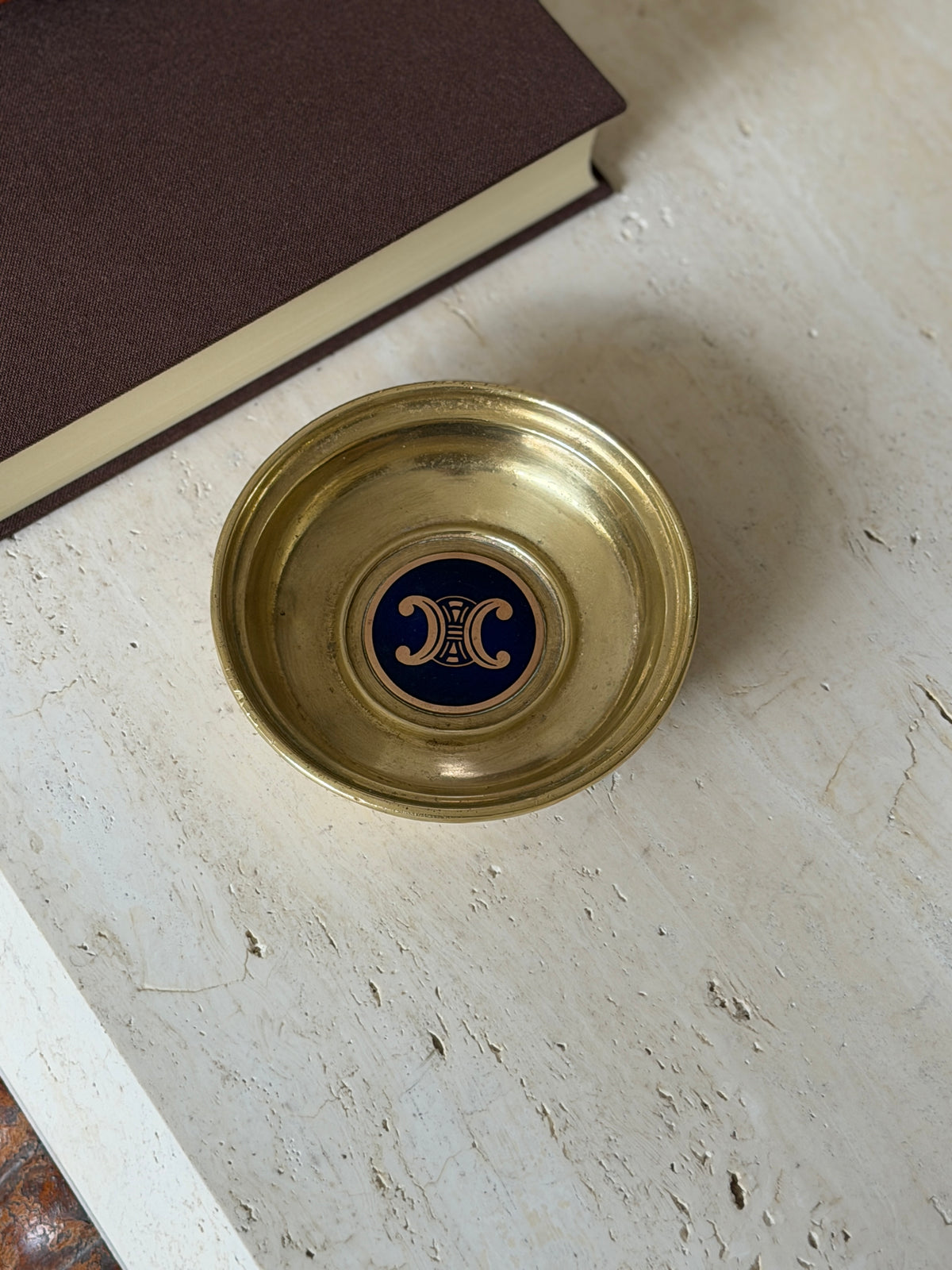 Overhead angled view of the brass dish, its center medallion in navy lacquer with the gold Triomphe emblem, resting on a stone surface near a book