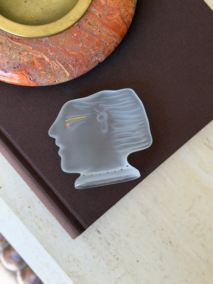 Overhead view of the Fassianos Kalisti crystal head lying on a brown book beside a red stone ashtray, the swept hair and ear molded in relief