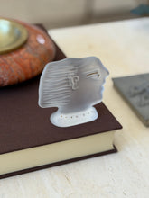 Frosted crystal profile head facing right on a brown book, showing the stylized ear, waved hair, and gold eye line