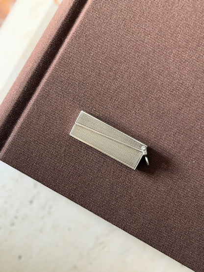 Dunhill solid silver travel cigar cutter closed, its engine turned barleycorn case and hinged suspension ring resting on a brown clothbound book