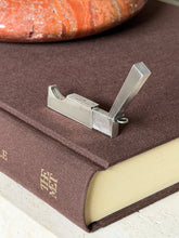 Open Dunhill silver cigar cutter standing on a brown book spine below a red stone ashtray, the lid raised at an angle