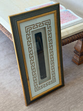 Angled view of the framed feng shui ruler leaning against a travertine table, an open art book resting on the tabletop above