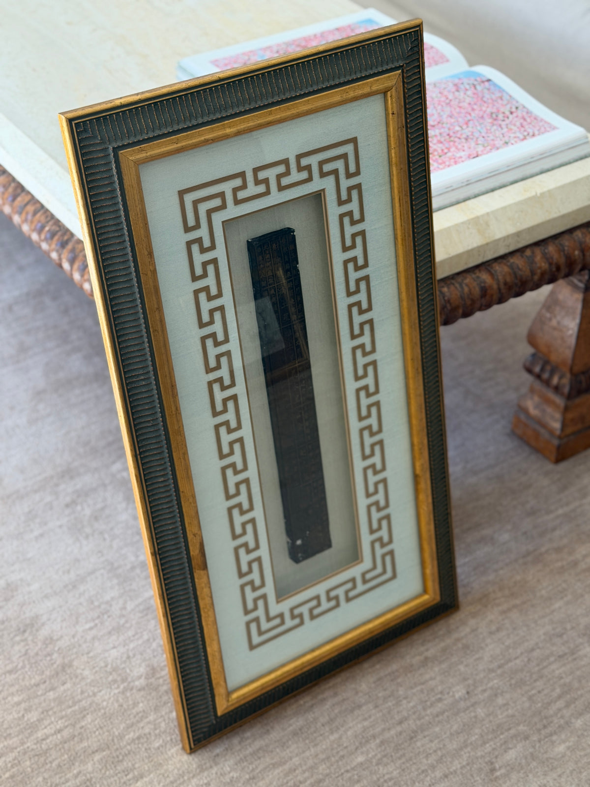 Angled view of the framed feng shui ruler leaning against a travertine table, an open art book resting on the tabletop above