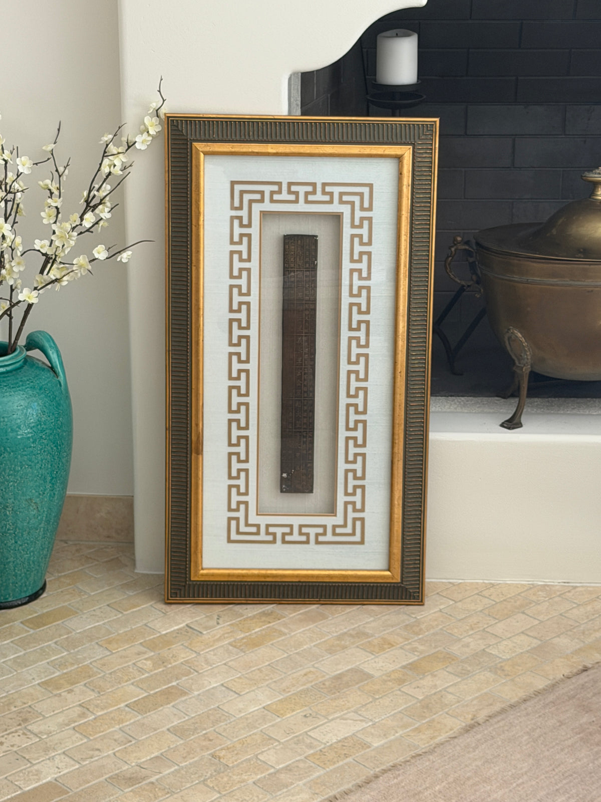 Front view of the framed Chinese feng shui ruler standing on a stone tile floor between a turquoise vase and a brick fireplace