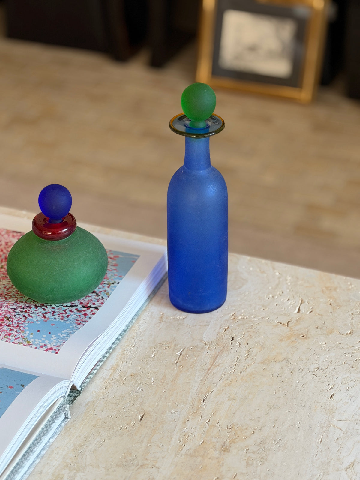 Tall cobalt blue scavo glass bottle with a frosted green ball stopper and flat amber-rimmed collar, standing at the edge of a travertine table
