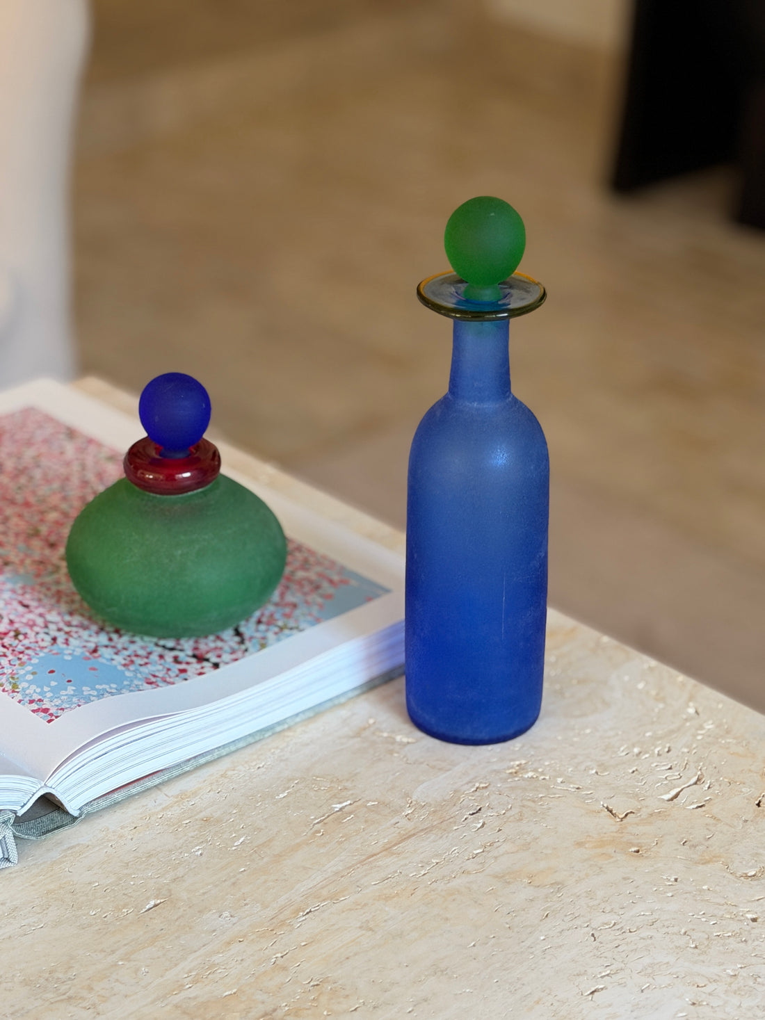 Close view of the Murano blue bottle showing its textured scavo surface and slender neck, a green stoppered bottle on an open book behind