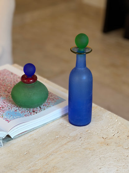 Close view of the Murano blue bottle showing its textured scavo surface and slender neck, a green stoppered bottle on an open book behind