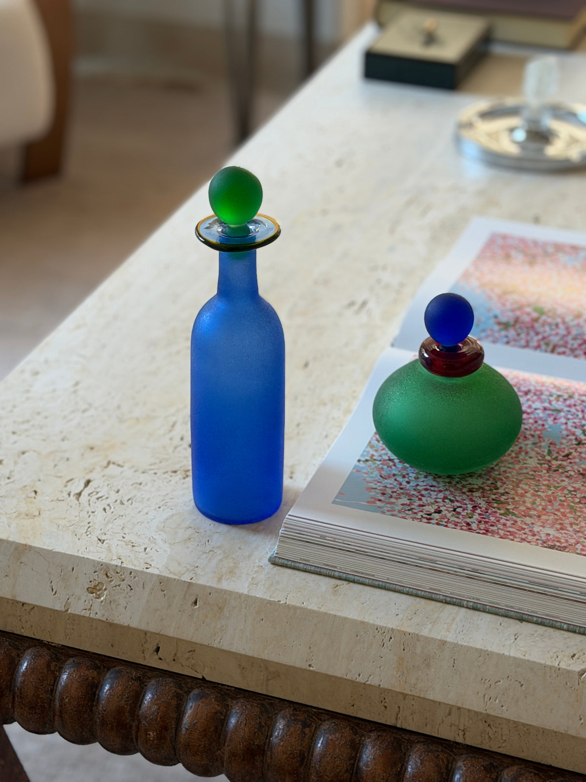 Blue scavo bottle on a travertine tabletop above a bobbin-turned wood base, the green vanity bottle resting on an open art book nearby