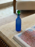 Franco Moretti blue glass bottle standing alone on travertine beside an open book of pink blossom prints, soft light on the frosted finish
