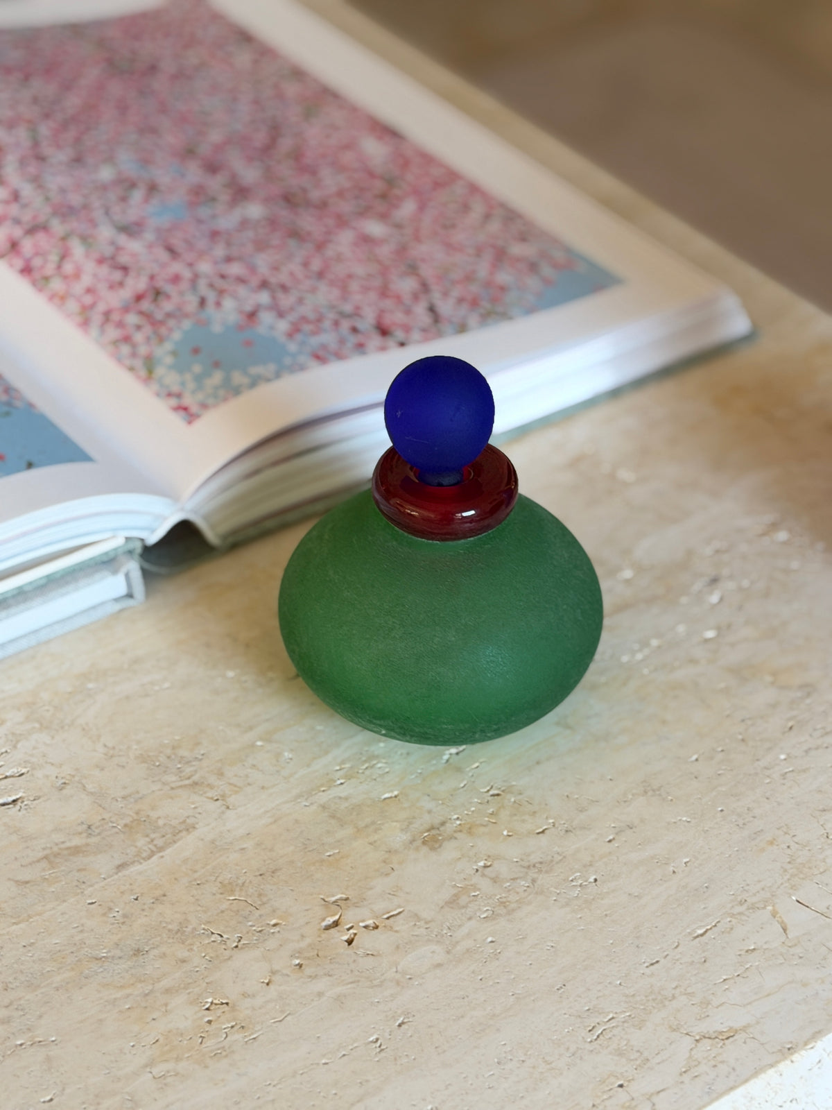 Squat green Murano scavo vanity bottle with a glossy ruby red collar and frosted cobalt ball stopper on a travertine table