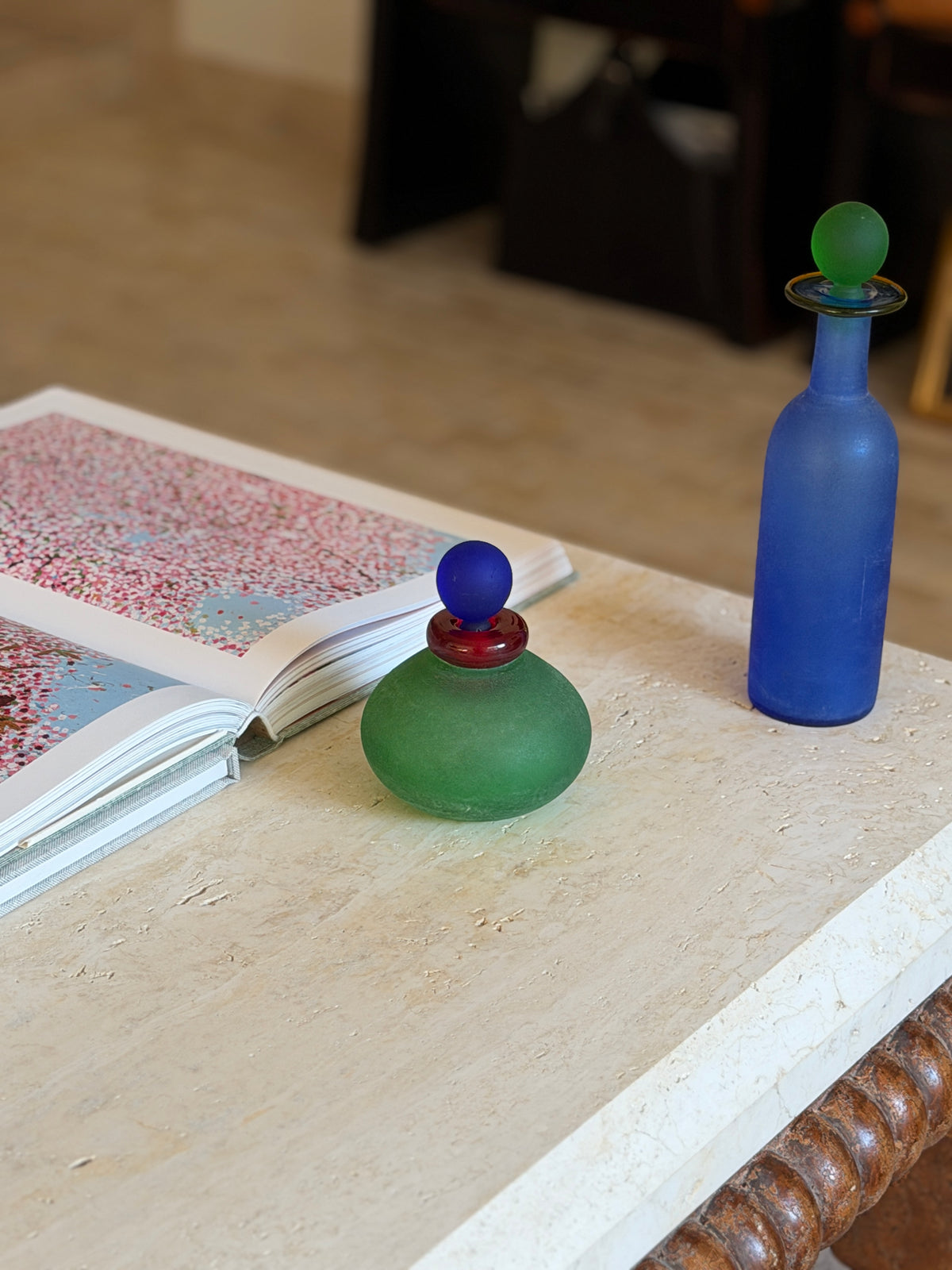 Green vanity bottle in front of an open art book with the tall blue scavo bottle standing near the table edge