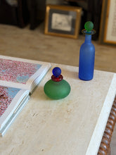Green Murano vanity bottle on travertine with the blue scavo bottle and gilt framed artwork softly blurred in the background