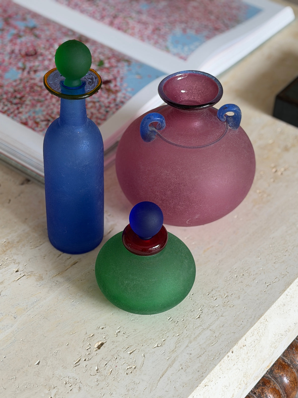 Franco Moretti pink scavo amphora vase with blue loop handles, shown with a blue bottle and green vanity bottle on travertine