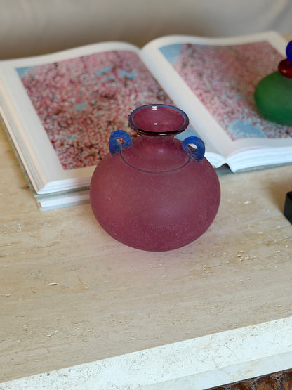 Raspberry pink Murano scavo amphora vase with two cobalt blue scroll handles resting in front of an open art book