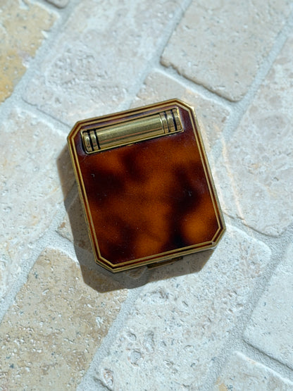 French Art Deco compact with a mottled tortoiseshell lid framed in brass, a banded brass lipstick tube set across the top, on tumbled stone pavers