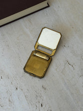 Open Art Deco compact on travertine, the mirrored lid raised above an engine turned brass powder lid and the black striped lipstick tube