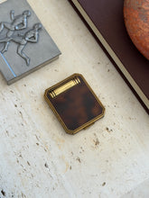 Closed tortoiseshell and brass compact on travertine beside a pewter box with athletes in relief and a red stone ashtray