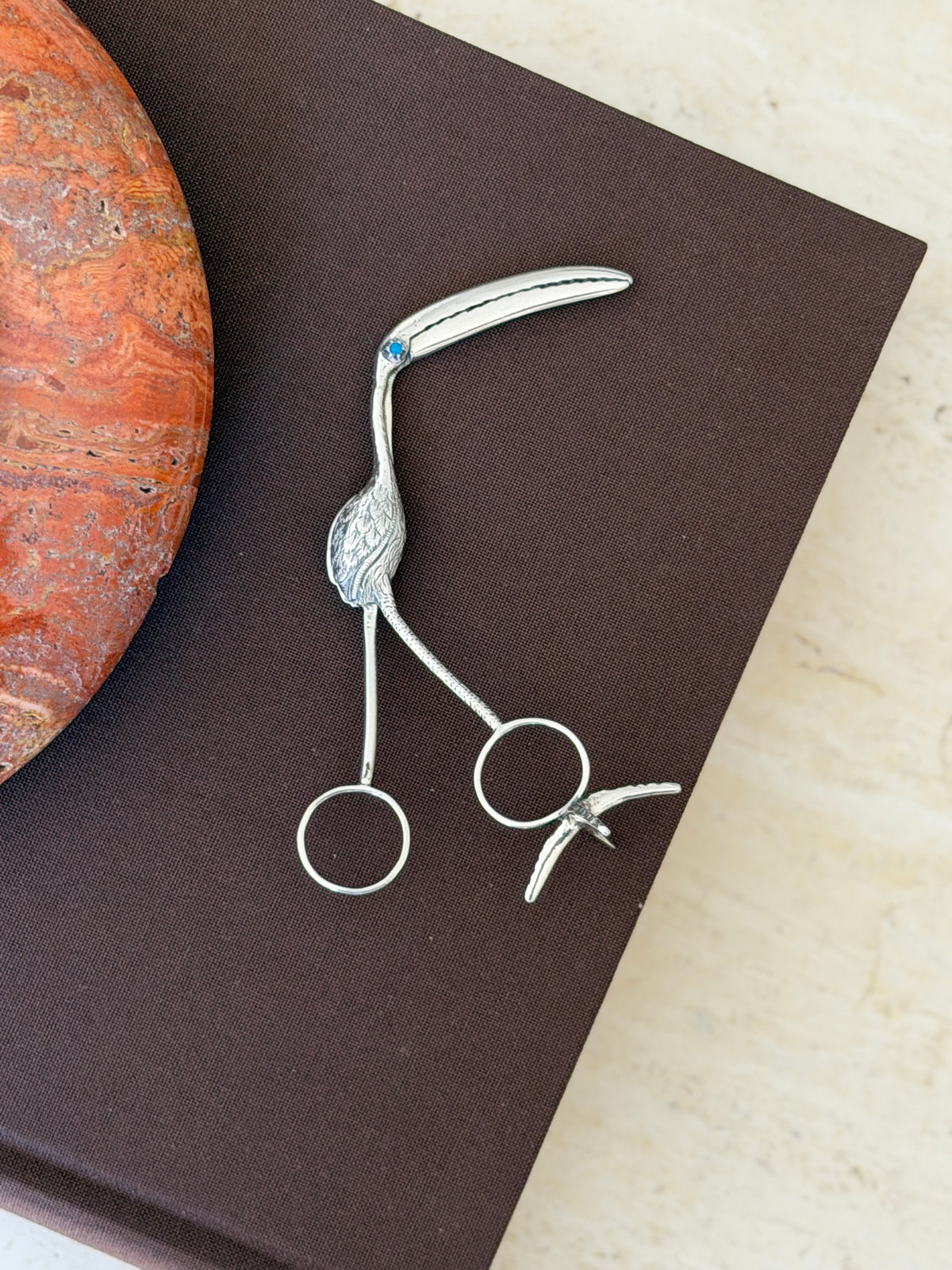 George Unite sterling silver flamingo sugar tongs with a long curved bill, engraved feathers, a turquoise eye, and ring handles, on a brown book