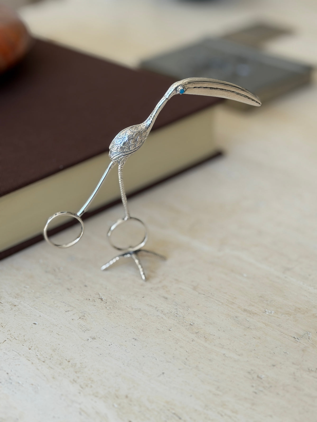 Silver flamingo tongs standing upright on their bird foot stand on travertine, the curved bill reaching forward