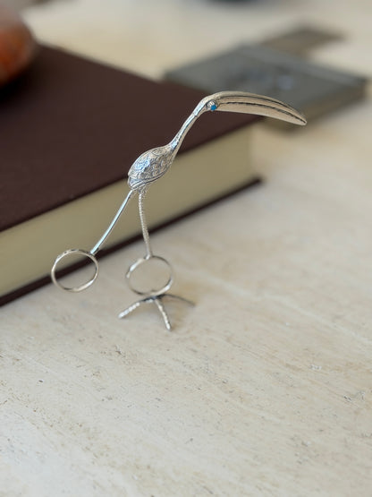 Silver flamingo tongs standing upright on their bird foot stand on travertine, the curved bill reaching forward