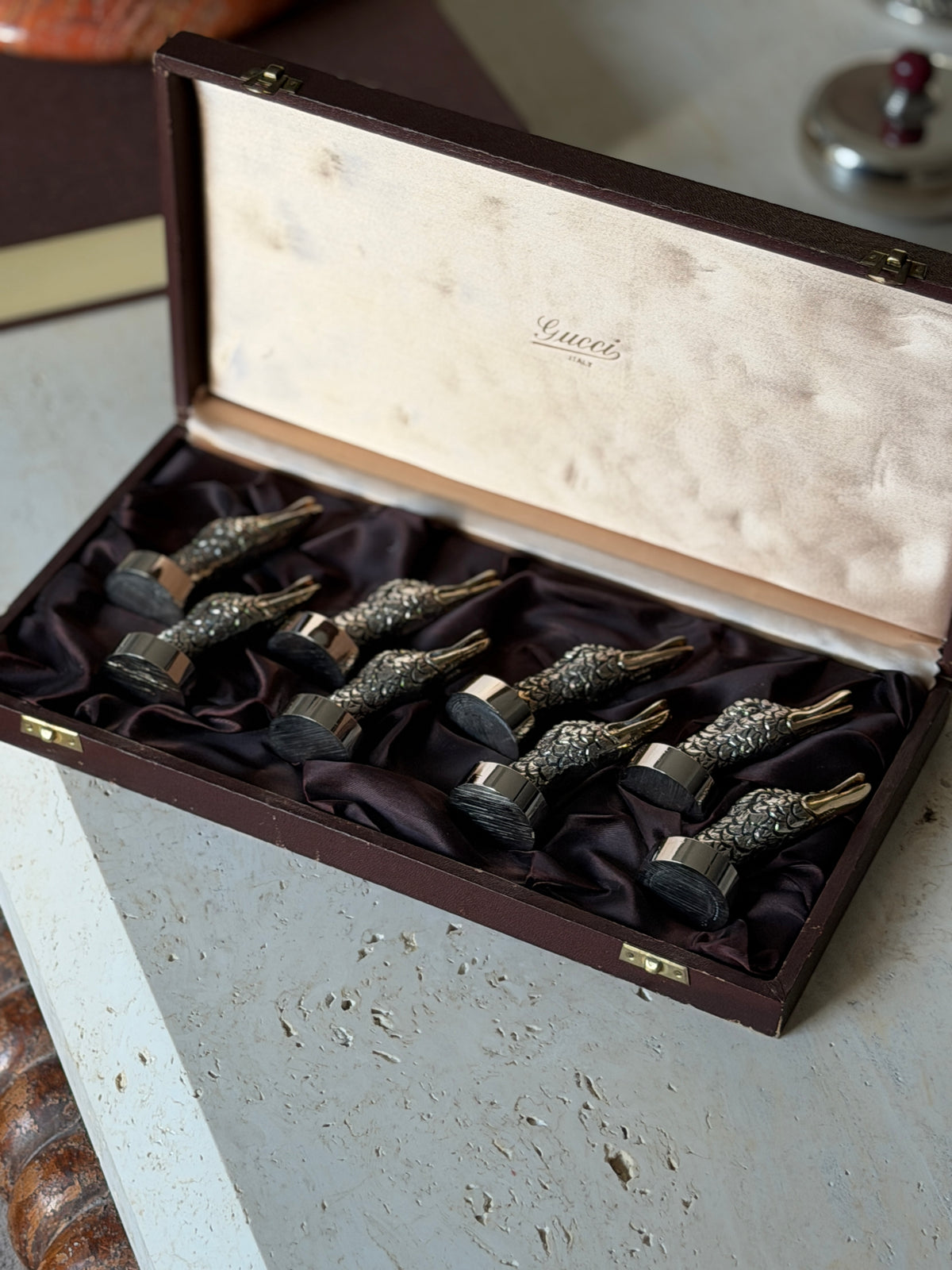 Set of eight Gucci duck head place card holders in their original burgundy presentation box, the cream satin lid stamped Gucci Italy, on a travertine table