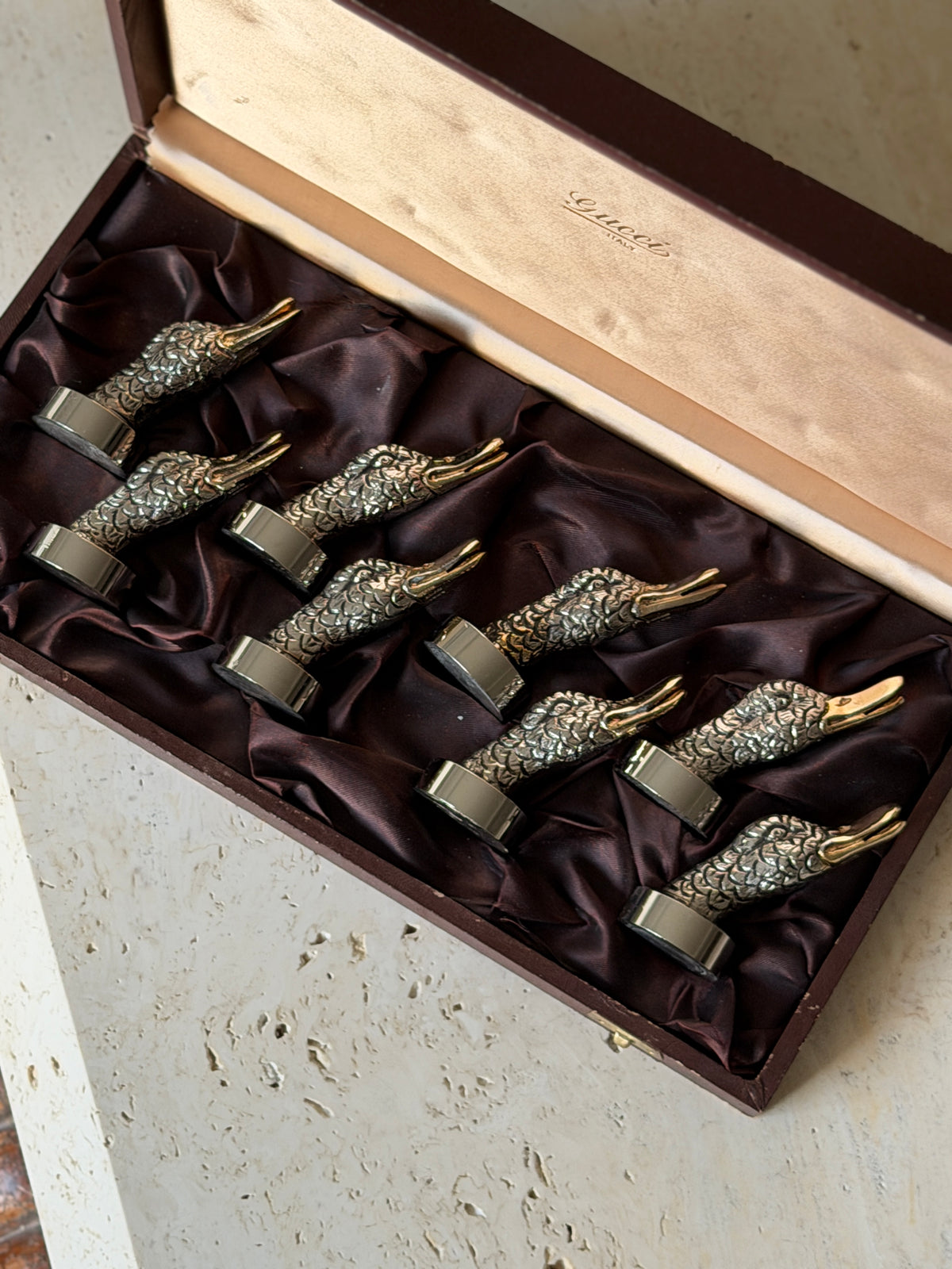 Overhead view of the Gucci place card holders nestled in plum satin, each silver tone duck head with finely chased feathers and a gilt beak on a round base