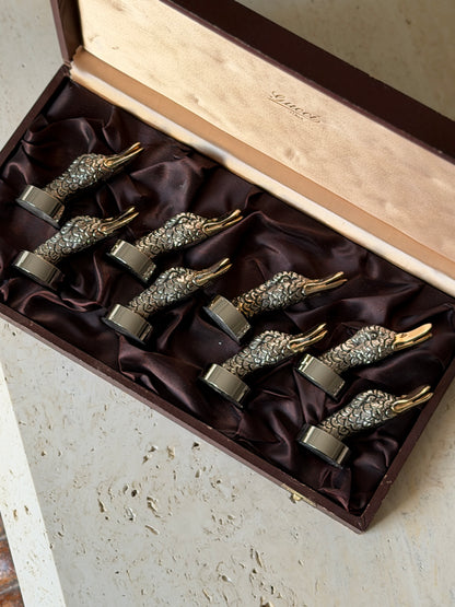 Overhead view of the Gucci place card holders nestled in plum satin, each silver tone duck head with finely chased feathers and a gilt beak on a round base