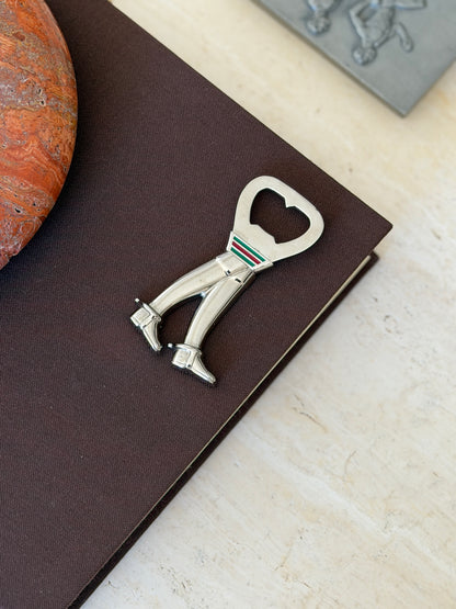 Gucci silver bottle opener shaped as a pair of riding boots with spurs, a green and red enamel stripe band at the top, on a brown book beside a red stone ashtray