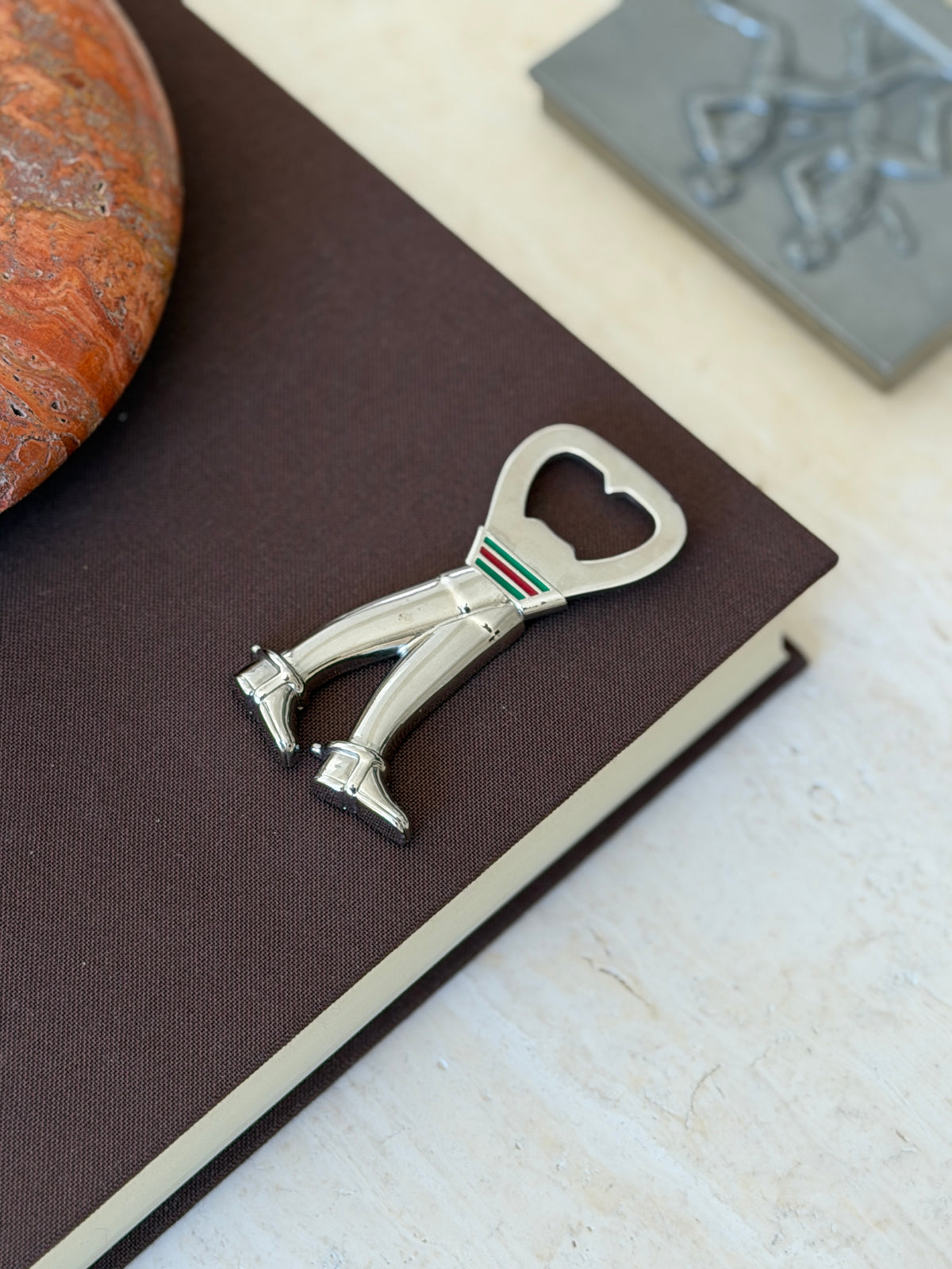 Angled view of the Gucci riding boot bottle opener on a brown book corner, the polished boots and buckled spur straps catching the light