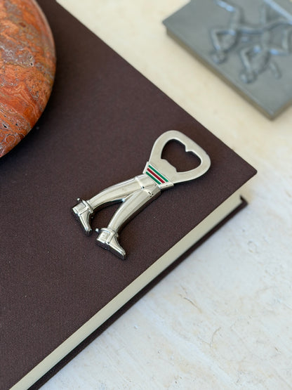 Angled view of the Gucci riding boot bottle opener on a brown book corner, the polished boots and buckled spur straps catching the light