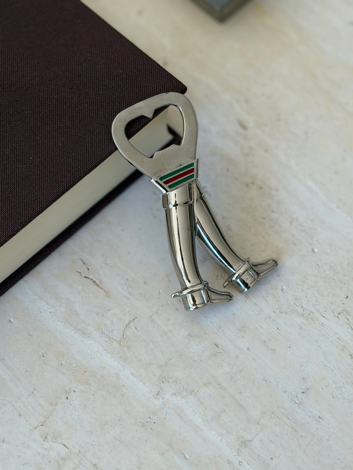 Gucci bottle opener leaning against a brown book on travertine, the boots standing side by side below the web stripe collar