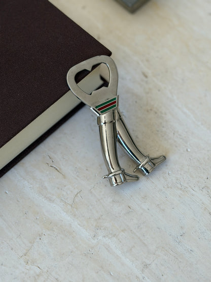 Gucci bottle opener leaning against a brown book on travertine, the boots standing side by side below the web stripe collar