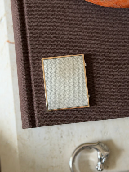 Closed folding frame resting on a fabric-bound book, showing the engine-turned silver panel and vermeil border