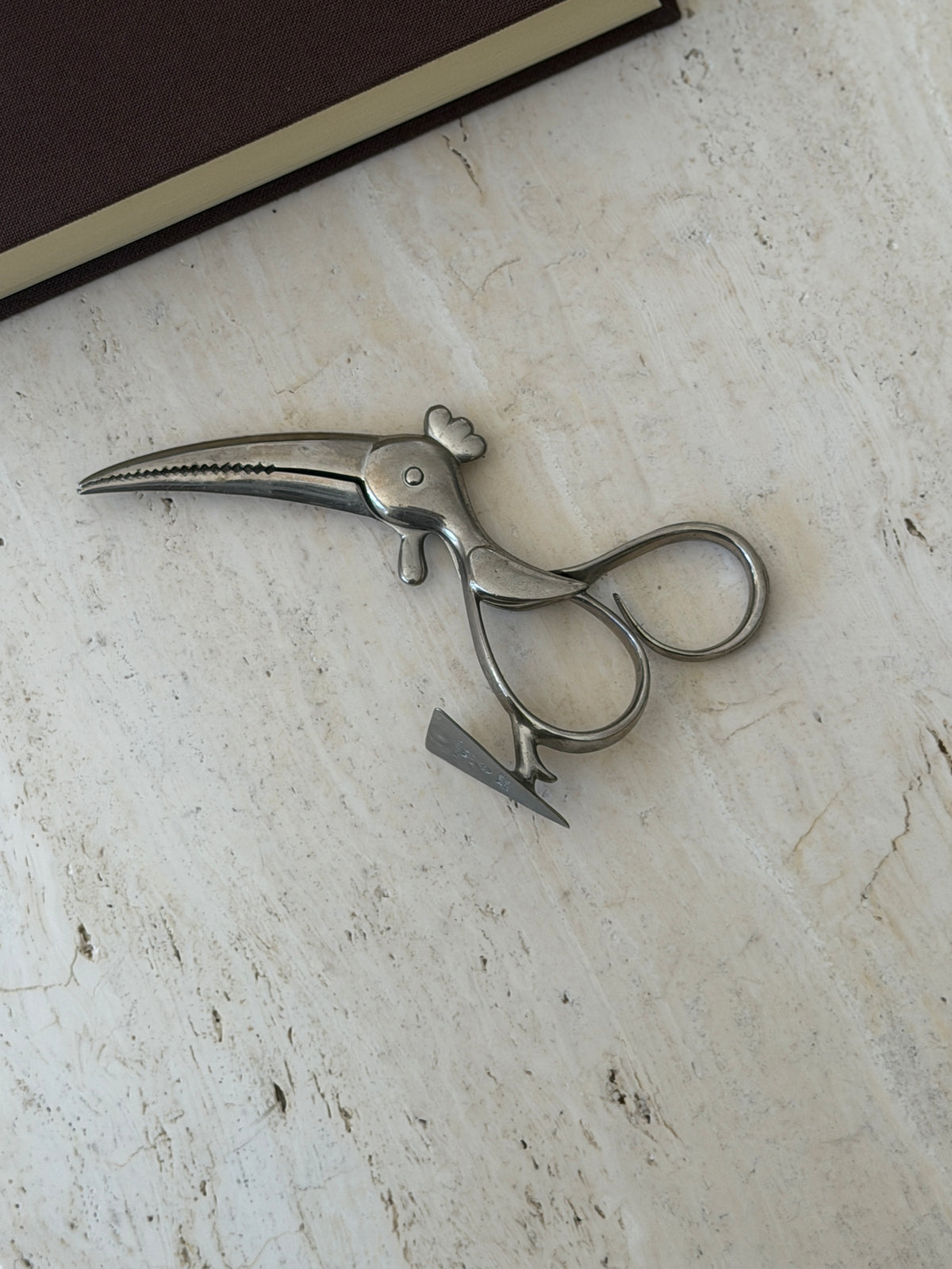 Hagenauer parrot sugar tongs lying on travertine, the long serrated beak forming the jaws and looped handles forming the body, beside a brown book