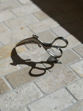 Silver-tone Hagenauer bird sugar tongs standing on a triangular foot on stone tile, raking sunlight casting a long sculptural shadow