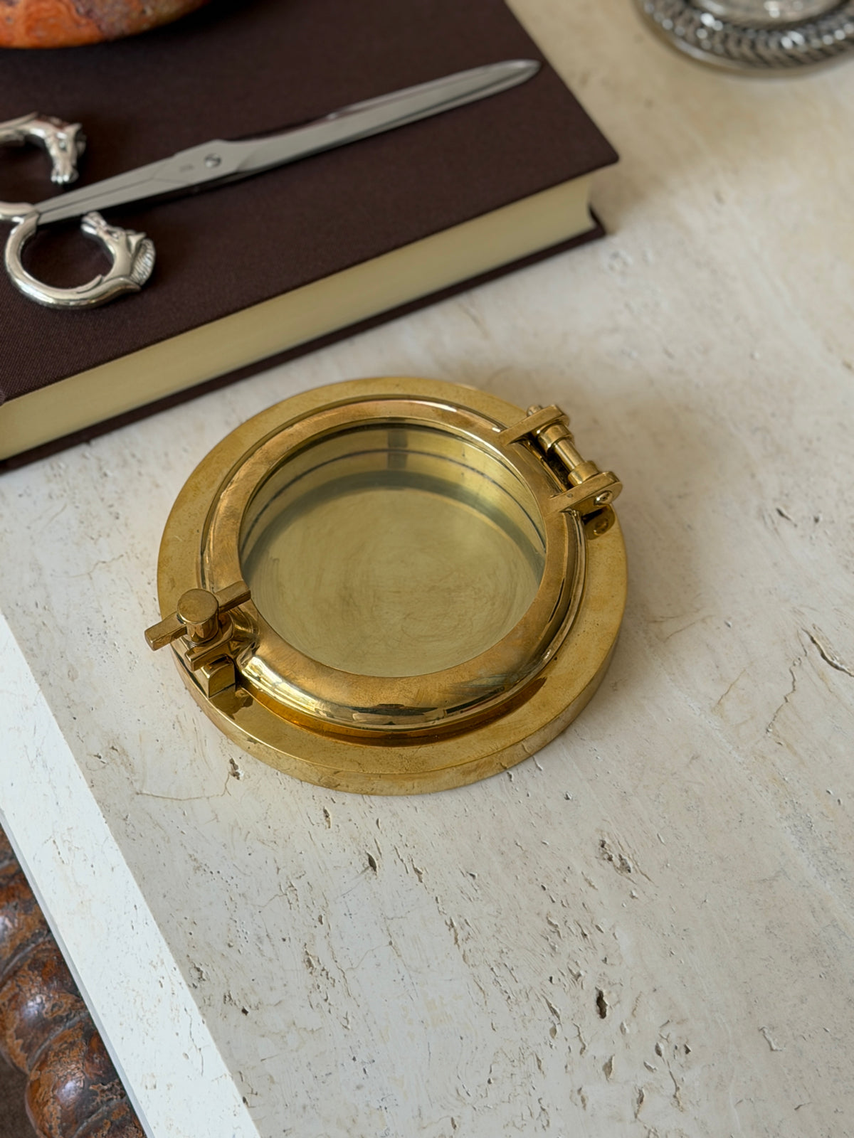 Overhead view of the closed brass porthole box with its wing nut latch, resting on a stone surface near an open book and a horse-head letter opener