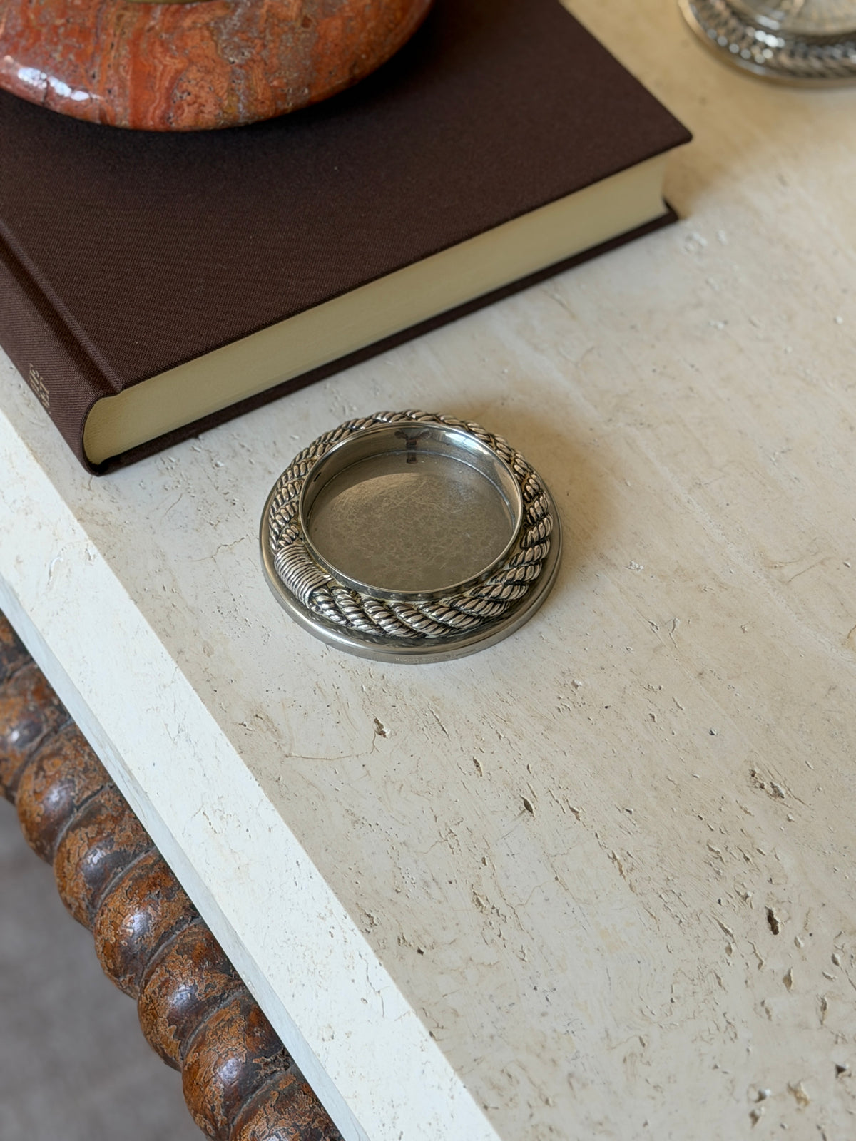 Hermes Cordage rope drink coaster in silver-toned metal on a travertine table beside a brown clothbound book, with stamped marks on the outer rim and inner wall