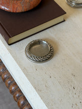 Hermes Cordage rope drink coaster in silver-toned metal on a travertine table beside a brown clothbound book, with stamped marks on the outer rim and inner wall