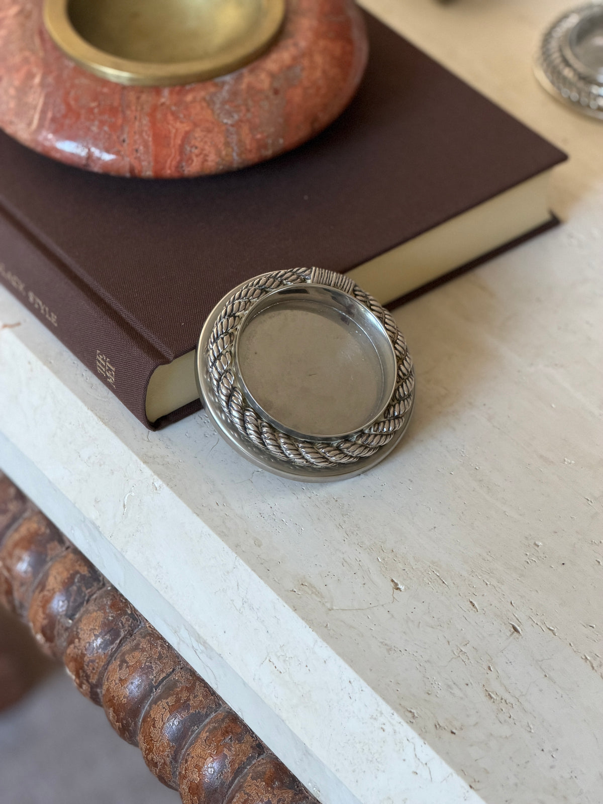 Hermes Cordage rope drink coaster propped on edge against a brown book, showing the depth of the rope border and polished outer rim, a red stone vessel behind