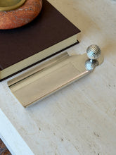 Hermes silver desk tray with an engine turned diamond pattern center and a dimpled golf ball mounted at one end, on travertine beside a brown book