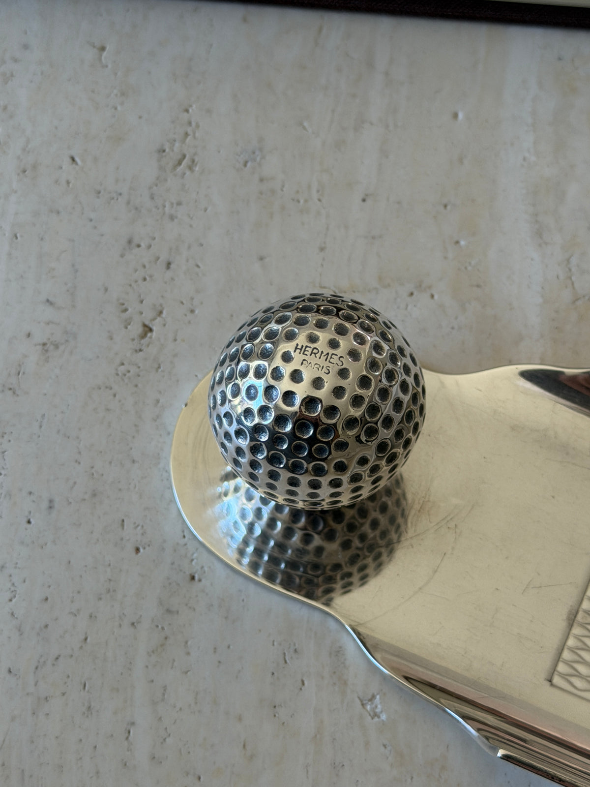 Close up of the dimpled silver golf ball on the Hermes tray, stamped HERMES PARIS on its top