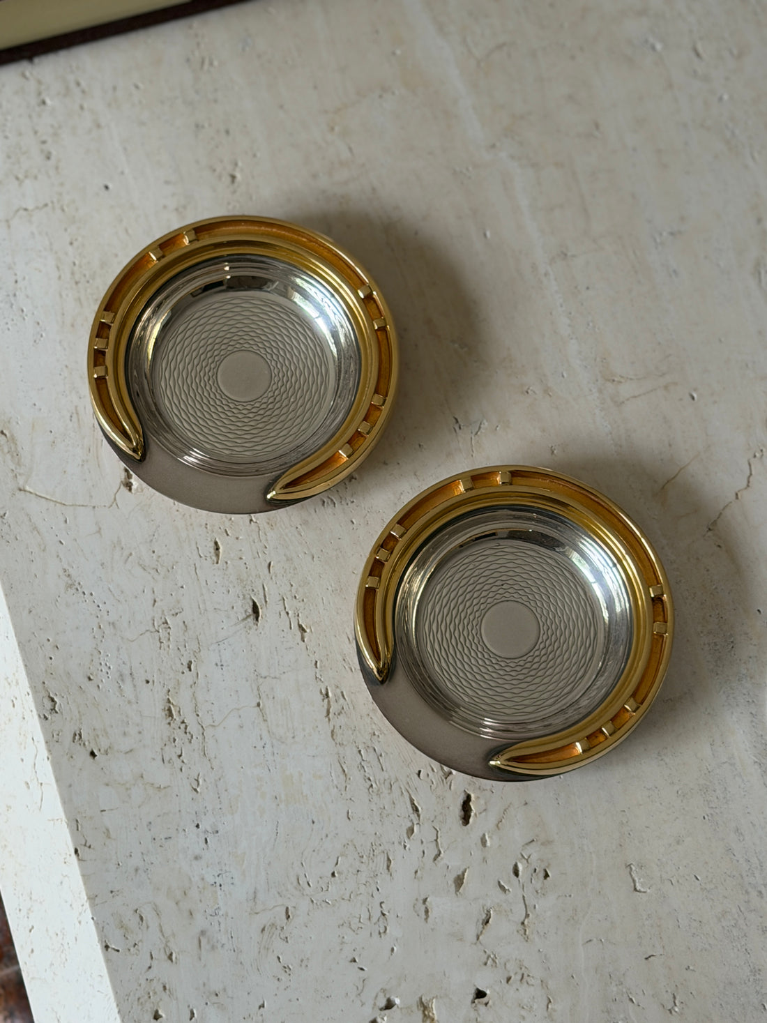 Two Hermes guilloche ashtrays with applied brass horseshoe rims shown from above on a travertine table