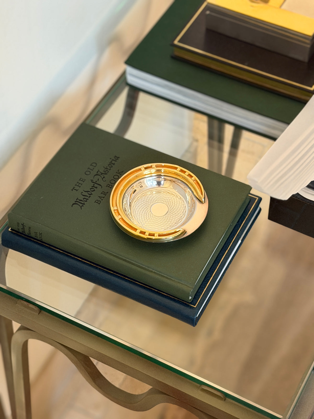 Hermes guilloche horseshoe ashtray resting on a green Old Waldorf-Astoria Bar Book on a glass-topped table